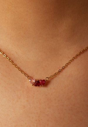 Collier en chaîne dorée avec trois pierres précieuses en forme de cœur : une rose et deux rouges. Design simple, reposant sur la peau avec une texture lisse.