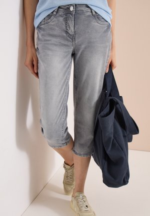 Person iført falmede grå cropped jeans, beige sneakers, lyseblå top og holder en stor marineblå tote taske på en lys baggrund.