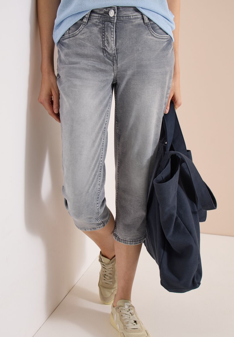 Personne portant un jean gris délavé raccourci, des baskets beiges, un haut bleu clair et tenant un grand sac cabas bleu marine sur un fond clair.