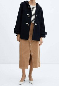 Cappotto navale sopra una camicia a righe, abbinato a una gonna di velluto a coste beige con bottoni frontali e décolleté bianchi a punta.