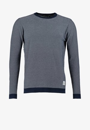 Homme marchant portant un pull gris, un jean foncé, des baskets bleues, et portant un sac noir sur une épaule.