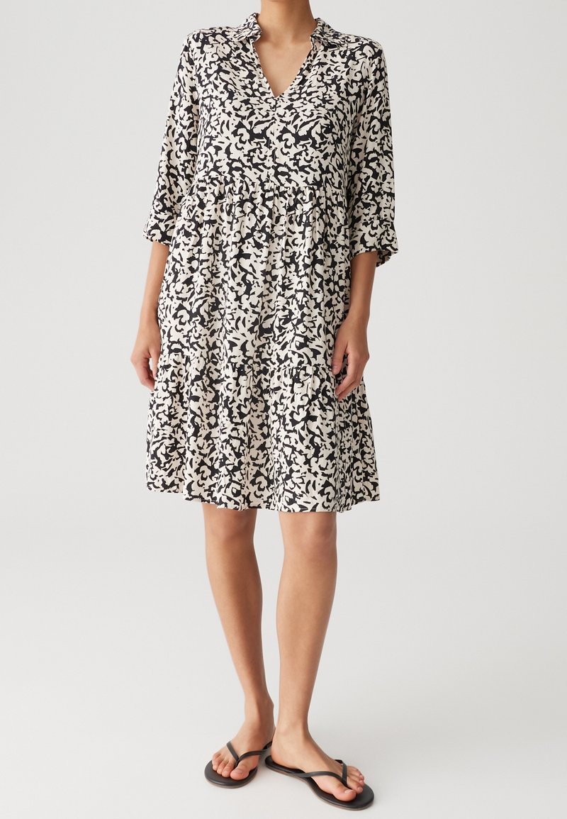 Femme portant une robe ample, mi-longue, à motifs noir et blanc, avec des manches trois-quarts et des sandales plates noires.