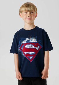 Niño con corte de pelo al bol, sonríe, vistiendo una camiseta de Superman azul marino con un gran logo y diseños gráficos sobre un fondo sencillo.