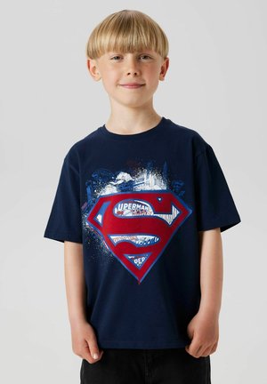 Un garçon avec une coupe de cheveux en bol sourit, portant un t-shirt Superman bleu marine avec un grand logo et des designs graphiques sur un fond uni.