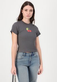 Top cropped cinza com nervuras e mangas curtas, apresentando um bordado de cereja no peito, combinado com jeans claros de cintura alta.