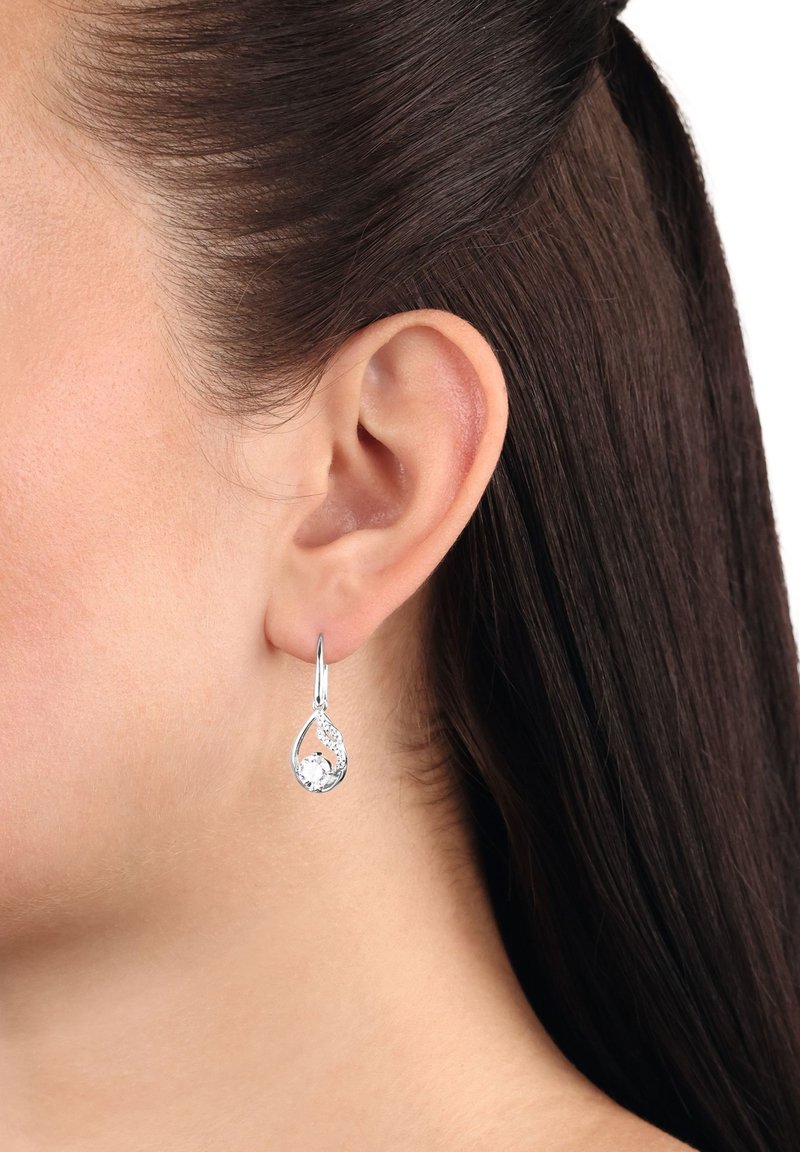 Närbild av en kvinnas öra med mörkbrunt hår som bär en silverfärgad droppformad örhänge med en rund, klar ädelsten.