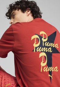 Camiseta de algodón roja con un gran diseño del logo de Puma en amarillo y negro en la parte posterior, con mangas cortas y un corte relajado.