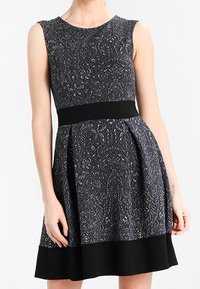 Schwarz-weiß gemustertes ärmelloses Kleid mit tailliertem Oberteil, schwingendem Rock und einfarbigem schwarzen Bund. Texturiert, knielang.