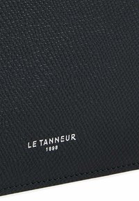 Black textured leather item featuring a subtle pattern, with a white embossed logo "LE TANNEUR 1898" near the bottom edge.