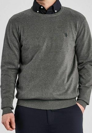Homme portant un sweat-shirt gris à col rond par-dessus une chemise noire boutonnée avec un pantalon foncé, main partiellement dans la poche.