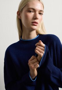 Marineblauw sweater met een zachte textuur, voorzien van een contrasterende lichtblauwe geribbelde halslijn en manchetten. Losvallend ontwerp, geen zichtbare patronen.