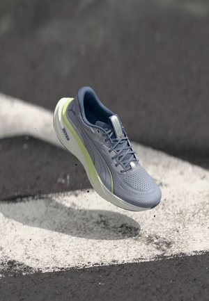 Scarpa da corsa grigia e giallo chiaro sospesa in aria sopra asfalto strutturato con segnaletica bianca e detriti sparsi.