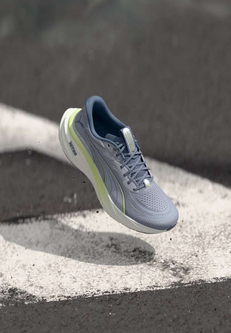 Γκρι και ανοιχτό κίτρινο αθλητικό παπούτσι στον αέρα πάνω από υφή άσφαλτου με λευκές διαγραμμίσεις δρόμου και διασκορπισμένα συντρίμμια.