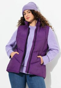 Lila Steppweste mit hohem Kragen, Reißverschluss und Seitentaschen, getragen über einem helllila Pullover. Das Model trägt eine passende Mütze.