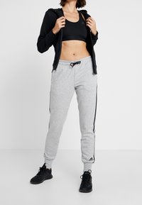 Sweat à capuche noir zippé porté sur un soutien-gorge de sport noir, associé à un pantalon de jogging gris avec des bandes latérales noires et des poignets élastiques, porté avec des baskets noires.