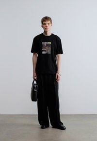 Μαύρο βαμβακερό T-shirt με σχέδιο στο μπροστινό μέρος, σε συνδυασμό με χαλαρά μαύρα παντελόνια. Κρατιέται μαύρη τσάντα στο δεξί χέρι.