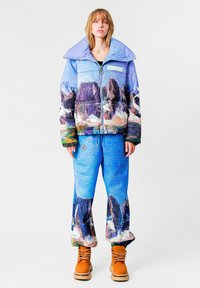 Chaqueta acolchada multicolor con un cuello ancho y estampado de paisaje. Los pantalones azules a juego presentan un diseño similar. Botas marrones completan el conjunto.