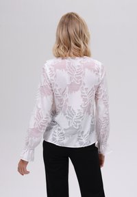 Hvit bluse med broderte bladmønstre, gjennomsiktig stoff, lange ermer med rynkede mansjetter, og rund nederkant, parret med svarte bukser.