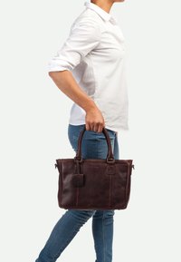 Sac à main en cuir marron avec deux anses, fermeture éclair et une étiquette de bagages. Texture lisse et forme structurée avec des coutures contrastantes.