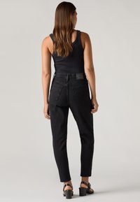 Mujer de pie, de espaldas, vistiendo una blusa negra sin mangas, jeans negros de cintura alta y sandalias negras de tacón, frente a un fondo liso.