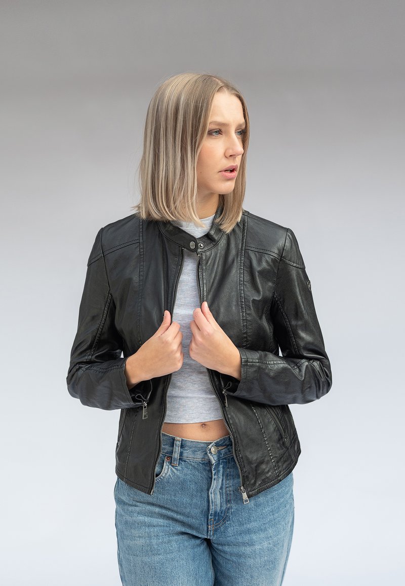 Junge Frau mit blonden Haaren, die eine schwarze Lederjacke, ein hellgraues Crop-Top und blaue Jeans trägt, hält den Jackenkragen und schaut zur Seite.
