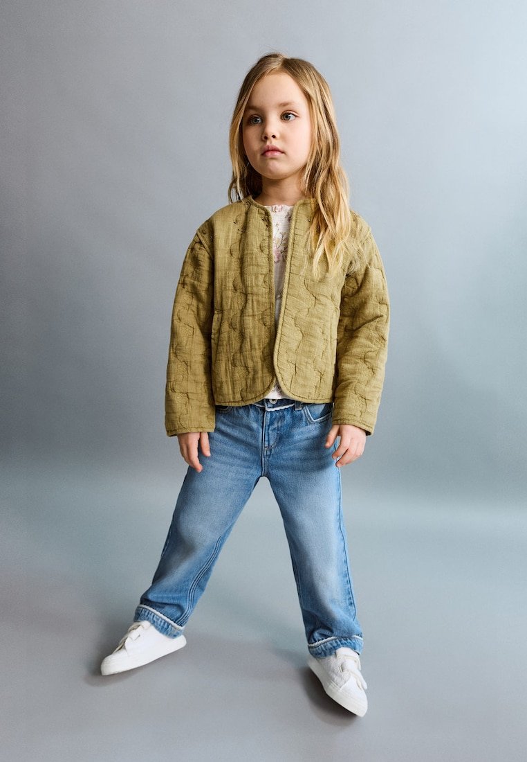 Jeune fille aux longs cheveux blonds portant une veste matelassée beige, un jeans bleu et des baskets blanches, debout devant un fond gris uni.