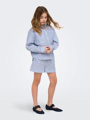 Noor tüdruk, kellel on pikad juuksed, seljas helesinise triibulise pikkade varrukatega särk, sobivate lühikeste pükstega ja mustad Mary Jane stiilis kingad, seisab valgel taustal.