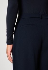 Personne portant un pantalon taille haute foncé avec une ceinture et un haut noir à manches longues, montrant la main avec une bague simple au doigt.