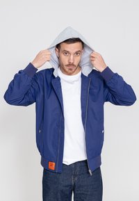 Blauwe bomberjack met grijze hoodievoering, rits aan de voorkant, elastische manchetten, twee zijzakken en een oranje label aan de zoom. Gestyled met een wit T-shirt.