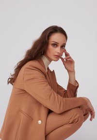 Karmelowy garnitur na miarę z dwurzędową marynarką i dopasowanymi spodniami. Marynarka ma złote guziki, a modelka nosi beżowy golf w prążki.