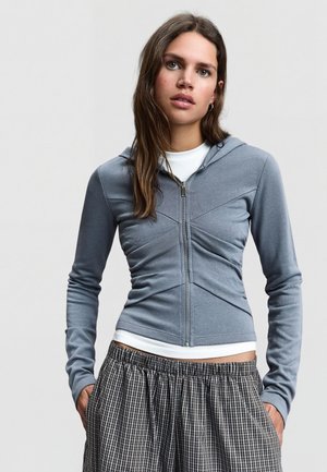 Jeune femme aux longs cheveux bruns portant un sweat à capuche gris ajusté zippé sur un t-shirt blanc et un pantalon ample à carreaux noirs et blancs.