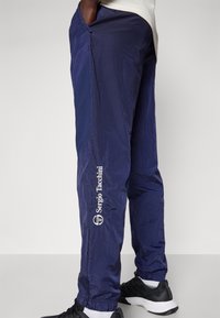 Námorné vetrové nohavice vyrobené z ľahkého materiálu, s elastickým pásom, bočnými vreckami a bielym logom na nohavici.