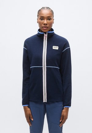 Temno modra flis jakna z visokim ovratnikom; ima roza zadrgo in modre poudarke ter logotip na levi strani prsnega koša.