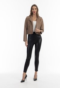 Giacca moto cropped in pelle sintetica marrone con dettagli metallici argentati, indossata sopra una maglietta bianca, abbinata a leggings neri a vita alta e tacchi neri.