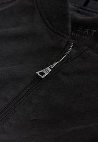 Gros plan d'une fermeture éclair partiellement ouverte d'une veste en daim noire, montrant le tire-zip en métal et le tissu doux texturé.