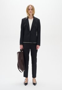Černý blazer na míru přes bílý top, doplněný úzkými černými kalhotami. Modelka drží hnědou koženou tašku a má na sobě černé špičaté baleríny.