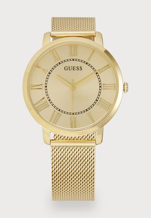 Arany színű óra minimalista kerek számlappal, római számokkal és hálós pánttal. A számlapon finom textúra található, valamint a "GUESS" márkanevével.
