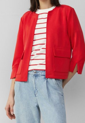 Kobieta ubrana w czerwoną krótka kurtkę z klapami i kieszeniami na kaniuki, na białej koszuli w czerwone paski oraz jasnoniebieskich, wysoko podniesionych dżinsach.