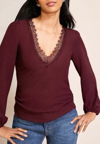 Burgunderfarbene Langarmbluse mit einem mit Spitze verzierten V-Ausschnitt, gerippter Textur und gerafftem Detail an der Taille, kombiniert mit blauen Jeans.