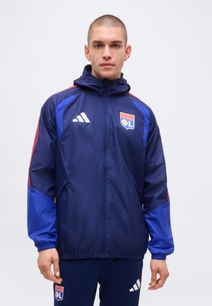 Uomo che indossa una giacca sportiva e pantaloni abbinati Olympique Lyonnais blu navy e rossi, in piedi contro uno sfondo semplice.