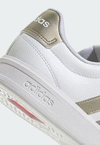 Zapatillas deportivas sintéticas blancas con detalles metálicos dorados, parte superior texturizada y una suela ligera que presenta un detalle rojo en la parte inferior.