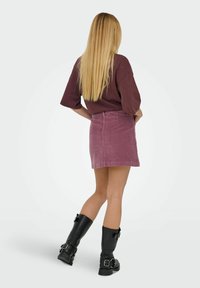 Mini-jupe en velours côtelé violet avec un ourlet brut, associée à une chemise marron ample et des bottes noires à genoux avec des accents de boucle.