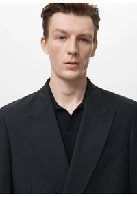 Black blazer with peaked lapels, textured black polo shirt underneath. Smooth fabric, fitted design, minimal detailing, and neutral backdrop.