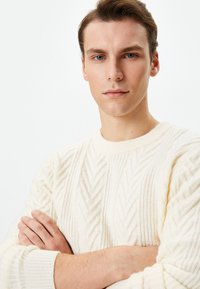 Suéter de punto color crema con un patrón de chevron texturizado. Cuello redondo y puños acanalados. Modelo de pie con los brazos cruzados.