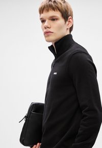 Pulôver preto com fecho na gola e logo no peito. O modelo segura uma bolsa de mão preta texturizada, que apresenta linhas limpas e um design minimalista.