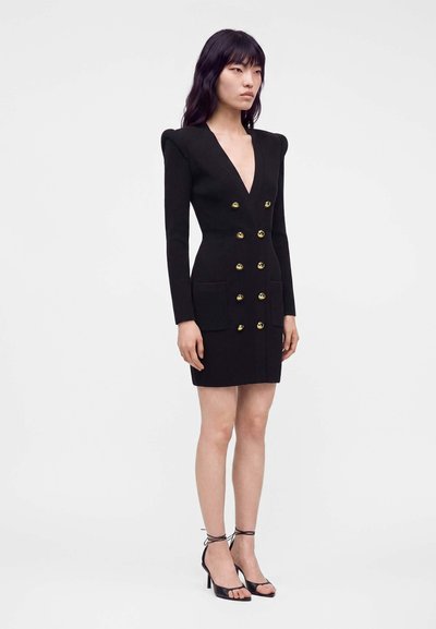 Femme debout portant une robe noire cintrée à double boutonnage avec des boutons dorés, des épaulettes, des poches frontales et des escarpins noirs à bride cheville.