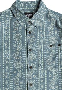 Korte mouwen overhemd met knopen in lichtblauw met paisley- en geometrische patronen. Heeft een borstzak en bruine knopen.