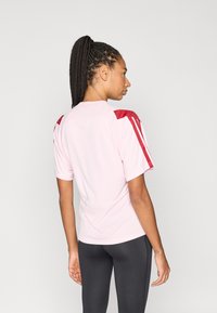 Világos rózsaszín sportos rövid ujjú póló piros válldíszítéssel. Légáteresztő anyagból készült, kerek nyakkivágással és karcsúsított fazonnal.