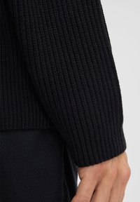 Manica di maglione nero a coste con una finitura testurizzata, con una bordatura a polsino aderente. Mano visibile che riposa vicino all'area del polso.