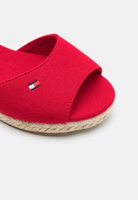 Sandalia tipo slip-on de lona roja con punta abierta, suela de cuerda de yute y parche pequeño con logo en colores azul, blanco y rojo.